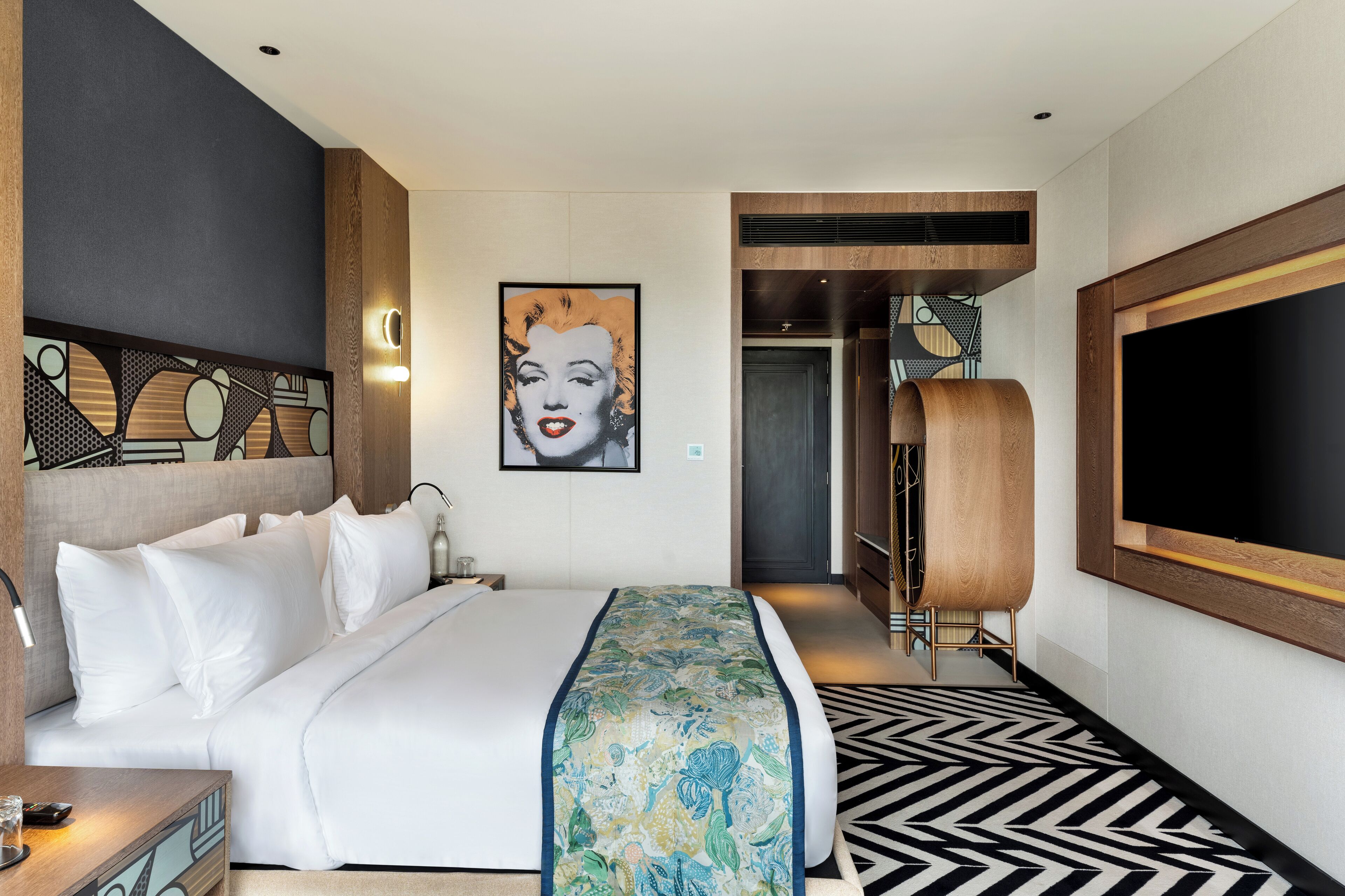 A hotel room with a large bed, a flat-screen TV, a decorative headboard, and a patterned rug.