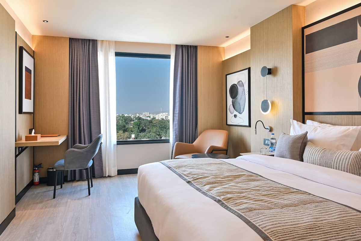 A hotel room with a large bed, a desk, a chair, and a view of a cityscape through the window.