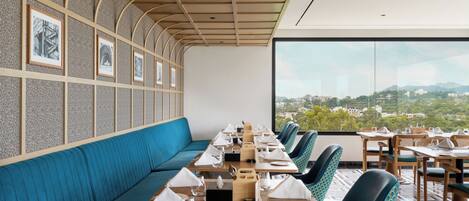 Een restaurant met een lange blauwe bank, houten tafels en stoelen, afbeeldingen in lijsten aan de muur en uitzicht op de stad.