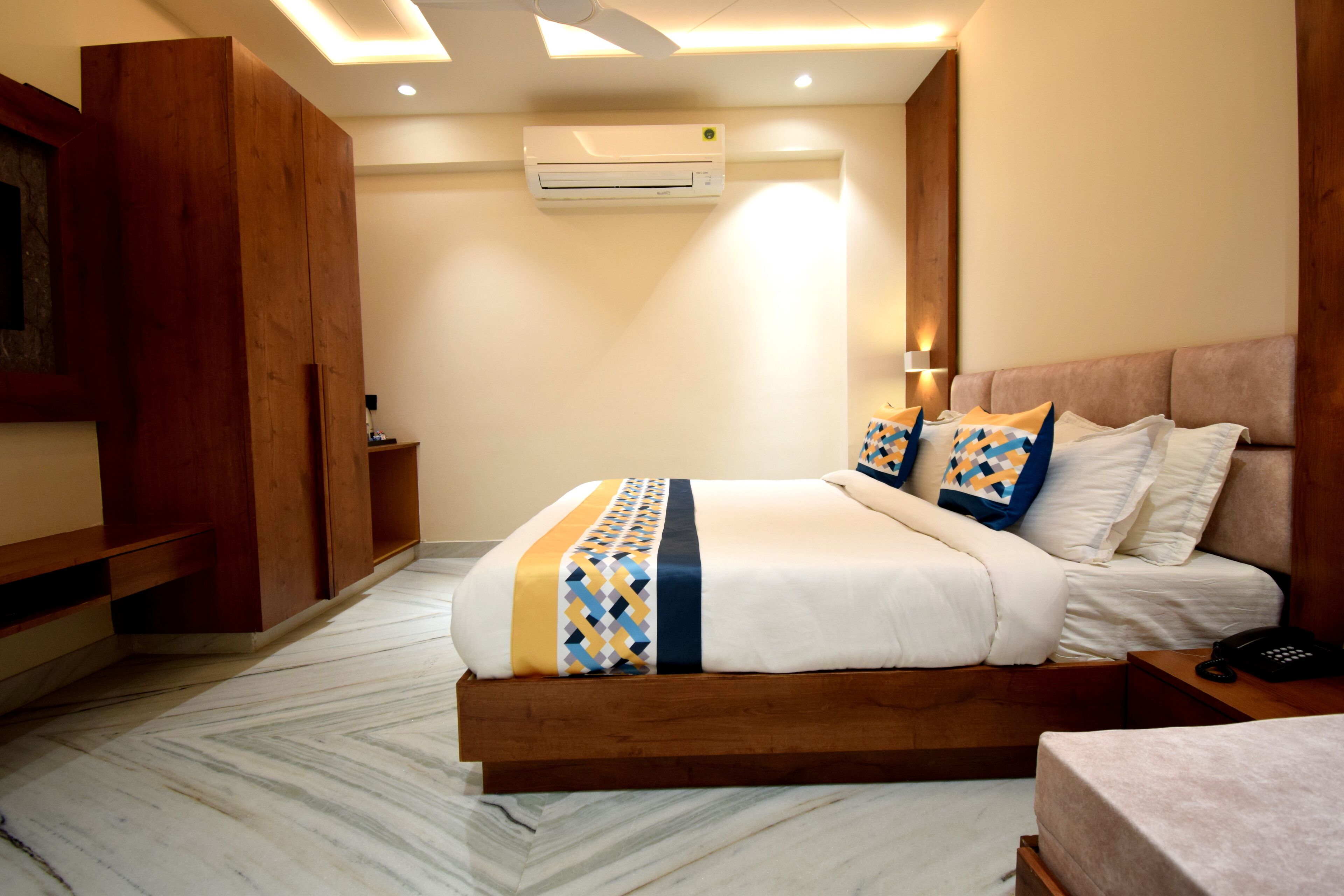 A hotel room with a wooden bed frame, a marble floor, a ceiling fan, an air conditioning unit, and a bedside table with a phone.