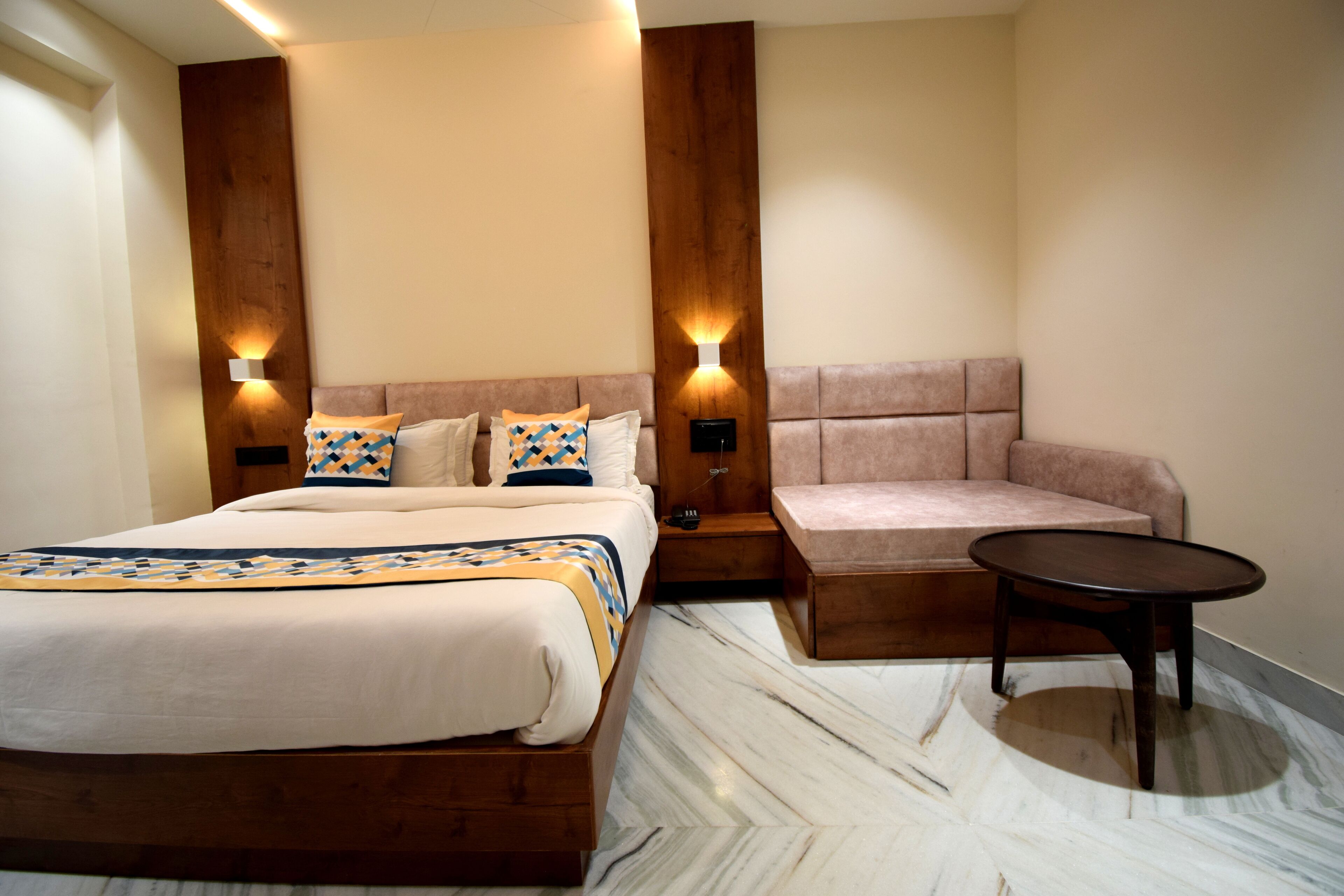 A modern hotel room with a large bed, a sofa, a small table, and two wall-mounted lights.