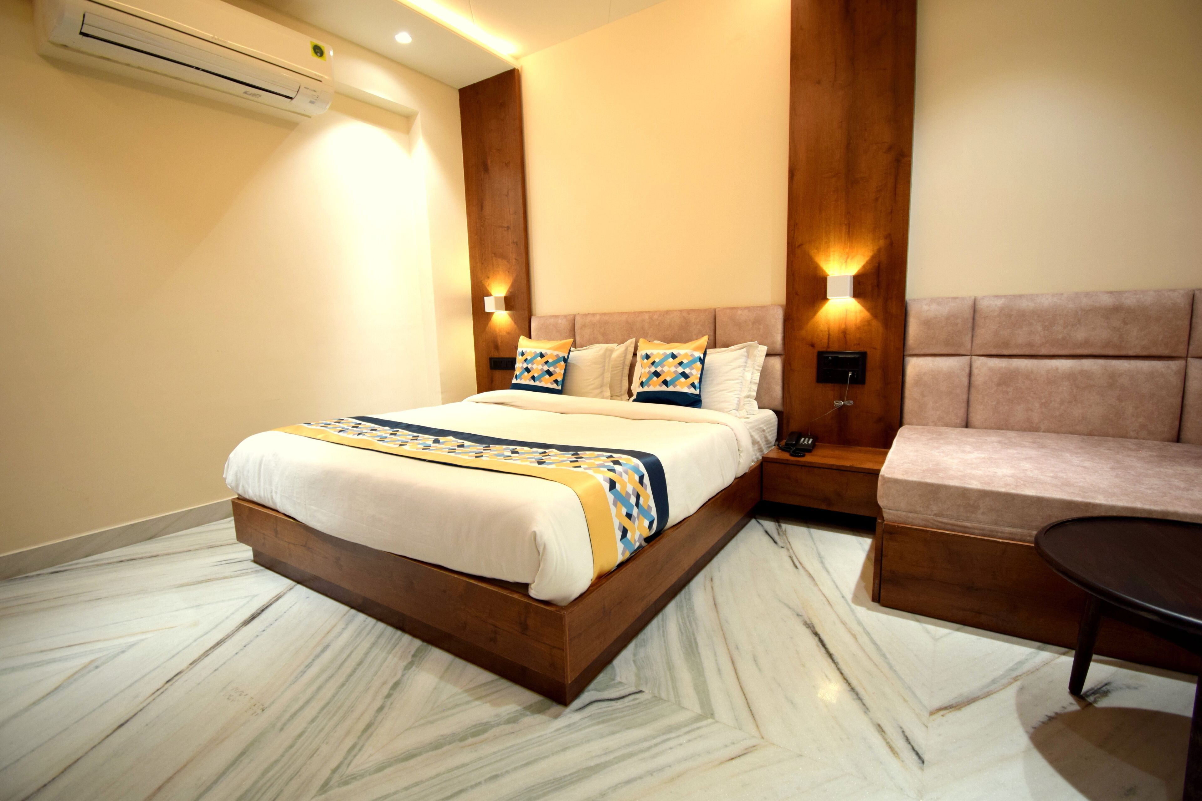 A hotel room with a large bed, a wooden headboard, a sofa, a small table, and a wall-mounted air conditioner.