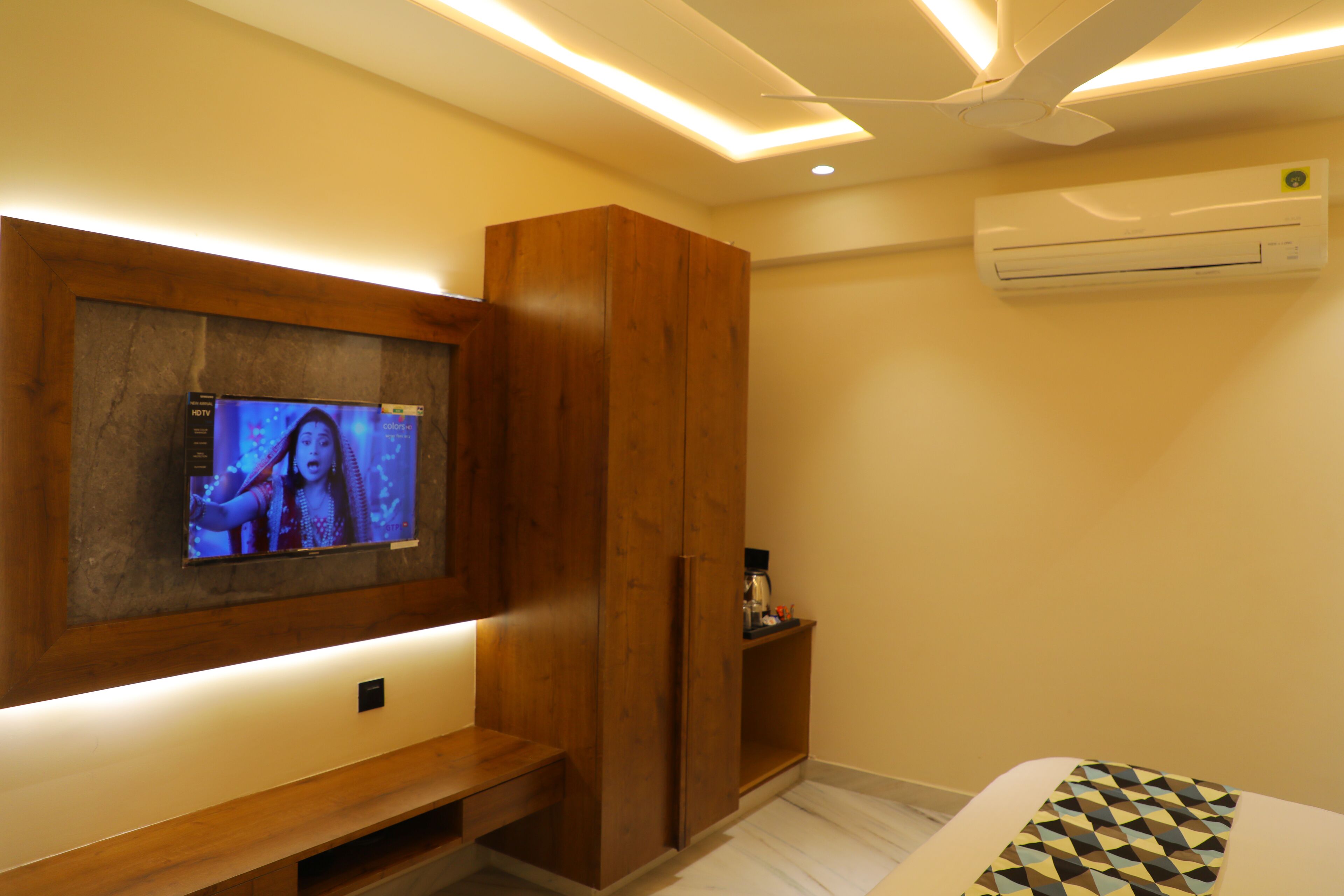 A hotel room with a TV mounted on the wall, a wooden wardrobe, a small shelf with items, and an air conditioning unit.