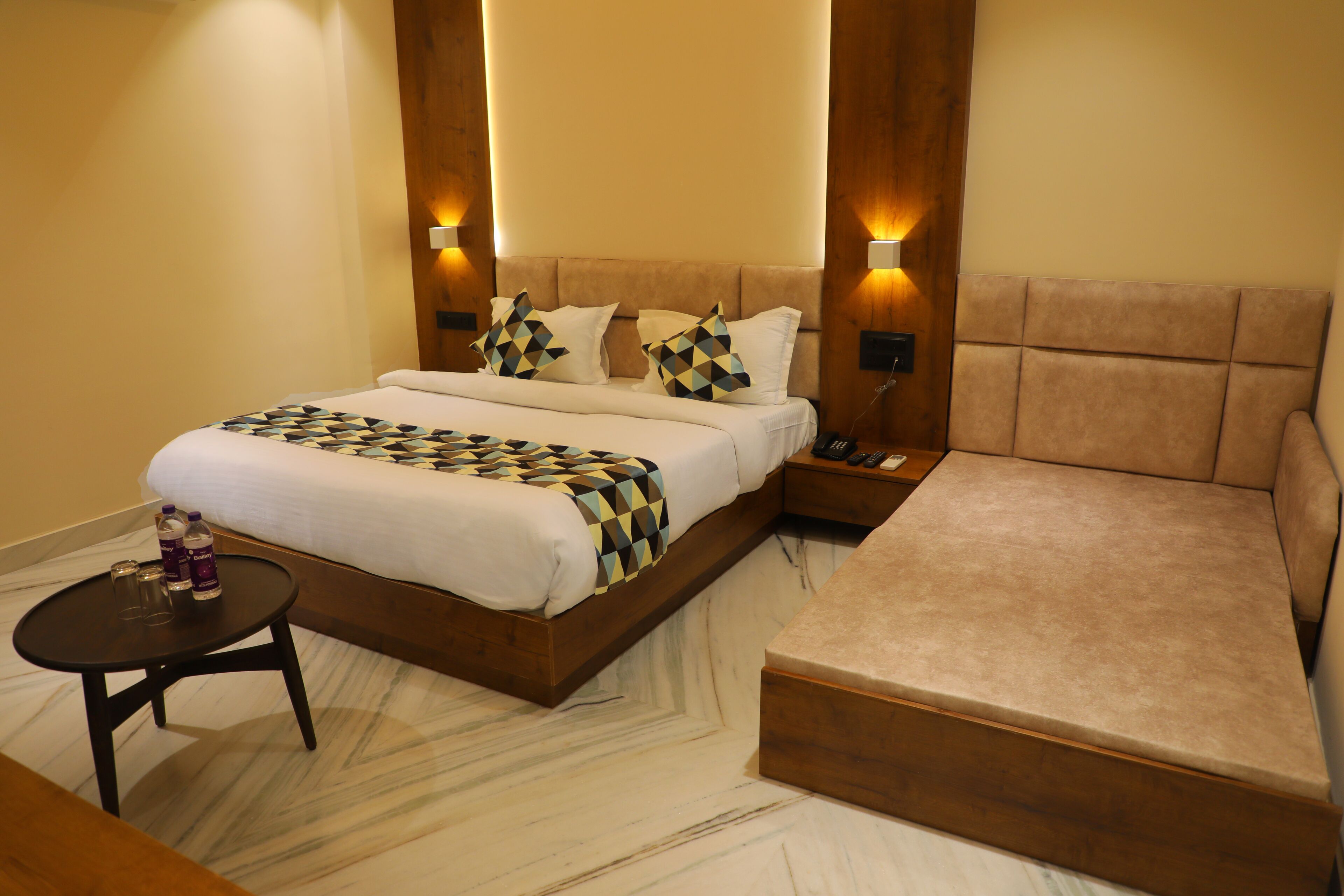 A modern hotel room with a large bed, a bedside table with a lamp, a small round table with bottles, and a sofa.