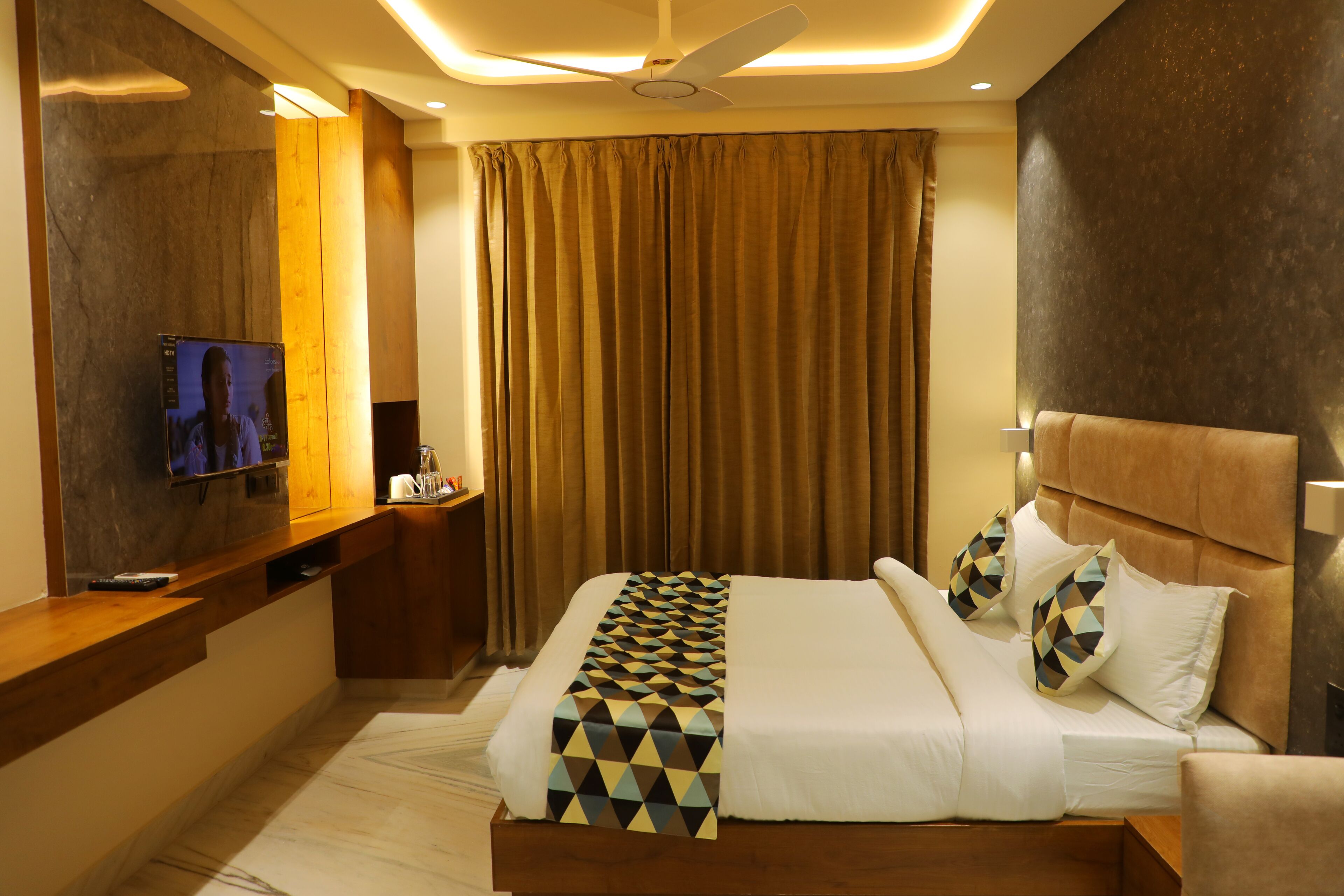 Une chambre d’hôtel moderne avec un grand lit, un téléviseur posé sur une console en bois et un ventilateur de plafond.
