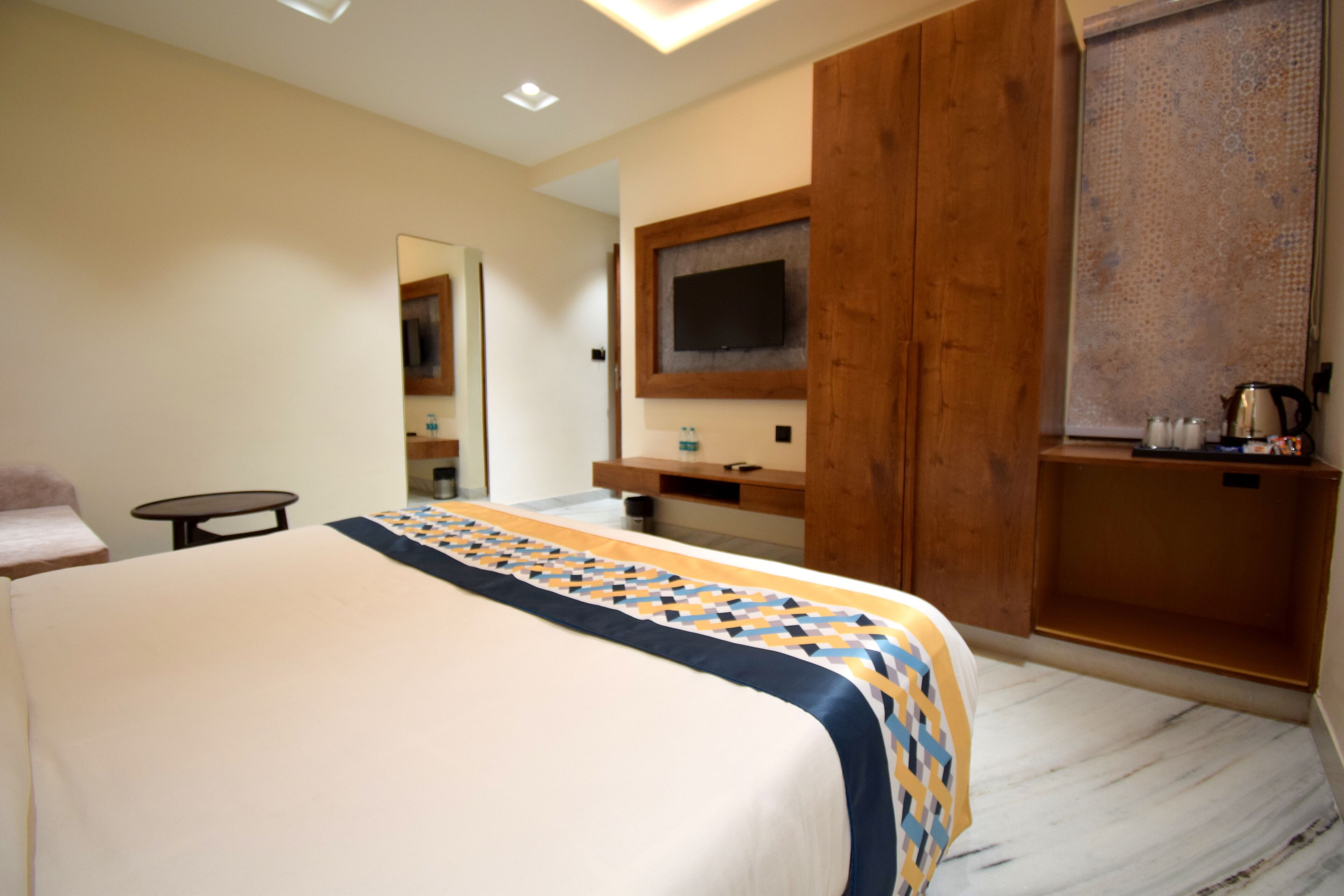 A hotel room with a large bed, a TV mounted on a wooden panel, a small table, and a bathroom area visible through an open door.