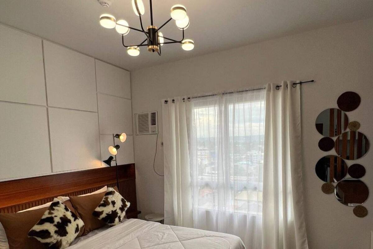 A bedroom with a bed, a headboard, a window with curtains, a wall-mounted mirror, and a ceiling light fixture.