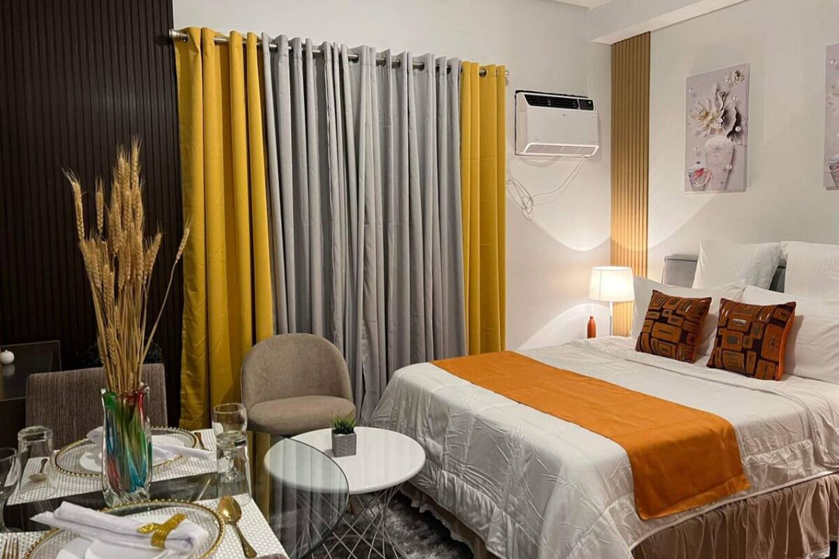 A hotel room with a bed, a chair, a small table, a vase with dried plants, and a wall-mounted air conditioner.