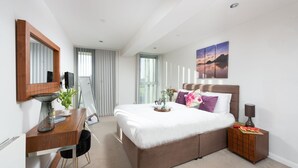 A modern hotel room with a large bed, a desk with a mirror, a bedside table with a lamp, and a view of the outdoors through the window.
