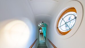 Una escalera estrecha con barandilla de madera, un ventanal redondo con vistas a edificios y un ventilador de techo.