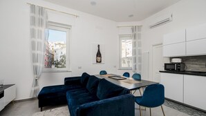 A modern living room with a dark blue sectional sofa, a dining table with chairs, and a kitchen area with white cabinets and a microwave. - Poerio 98 Luxury Apartment (Naples)