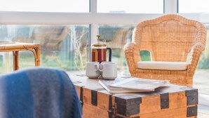 Un coin salon accueillant avec un fauteuil en osier et une table en bois, une valise ancienne et une cafetière.