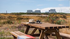 Une table de pique-nique en bois, sur laquelle sont posés un chapeau, des jumelles et un livre, est installée dans un champ herbeux, avec un bâtiment en arrière-plan.