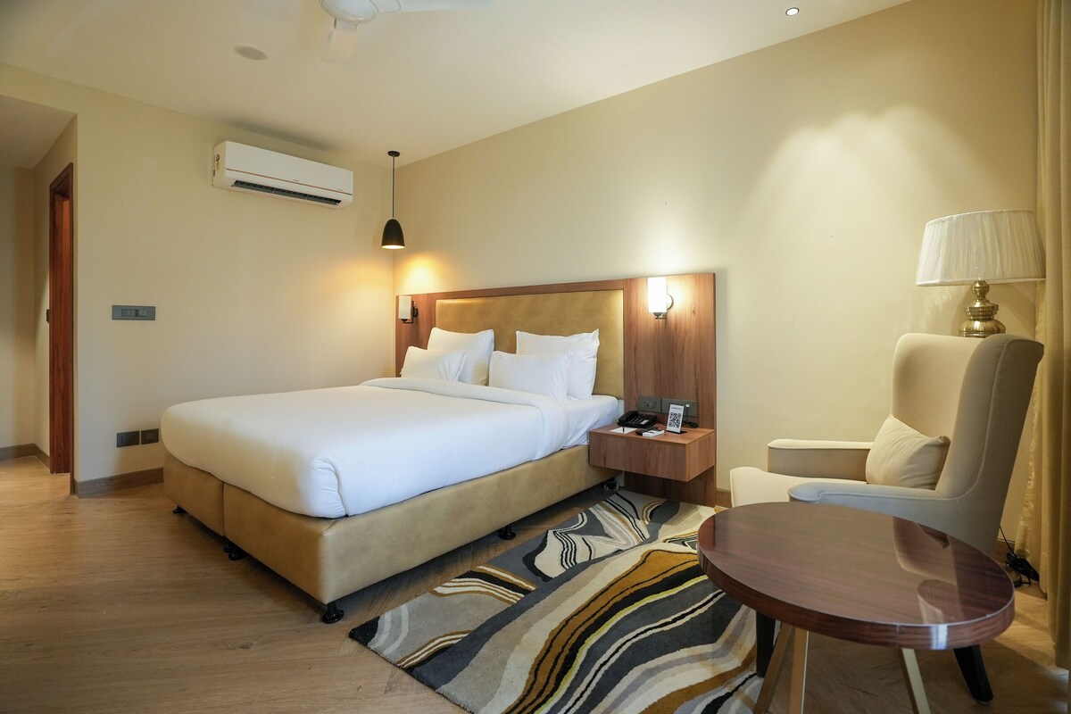 A hotel room with a large bed, a bedside table with a lamp, a chair, and a small table.