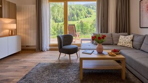 Un salon moderne avec un canapé, un fauteuil et une table basse. Une grande fenêtre offre une vue sur une zone boisée.