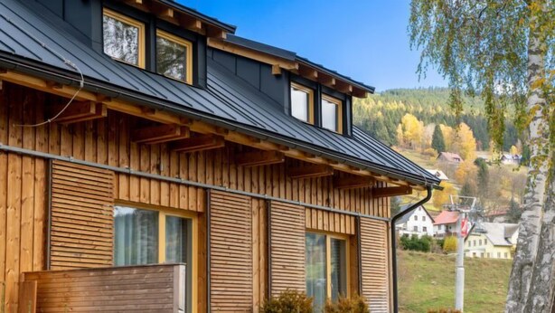 Une maison moderne en bois, dotée d’un toit à pente, de grandes fenêtres et offrant une vue sur une colline boisée.