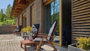 Une terrasse extérieure aménagée avec du mobilier en bois, comprenant des chaises et une table, et une grande fenêtre donnant sur un paysage verdoyant.