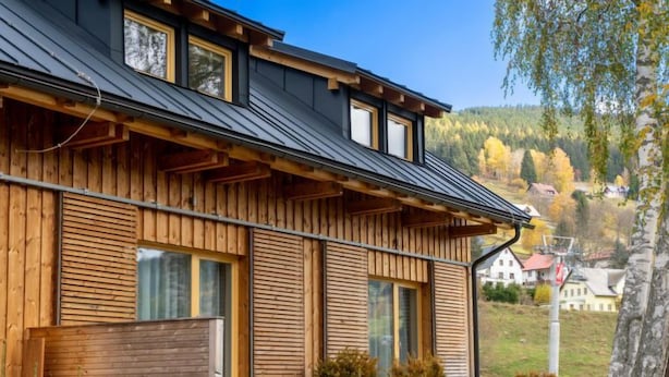 A modern wooden house with a sloped roof, large windows, and a view of a forested hillside.