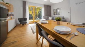 A modern kitchen with a dining area, wooden flooring, and a large window with a view of trees.