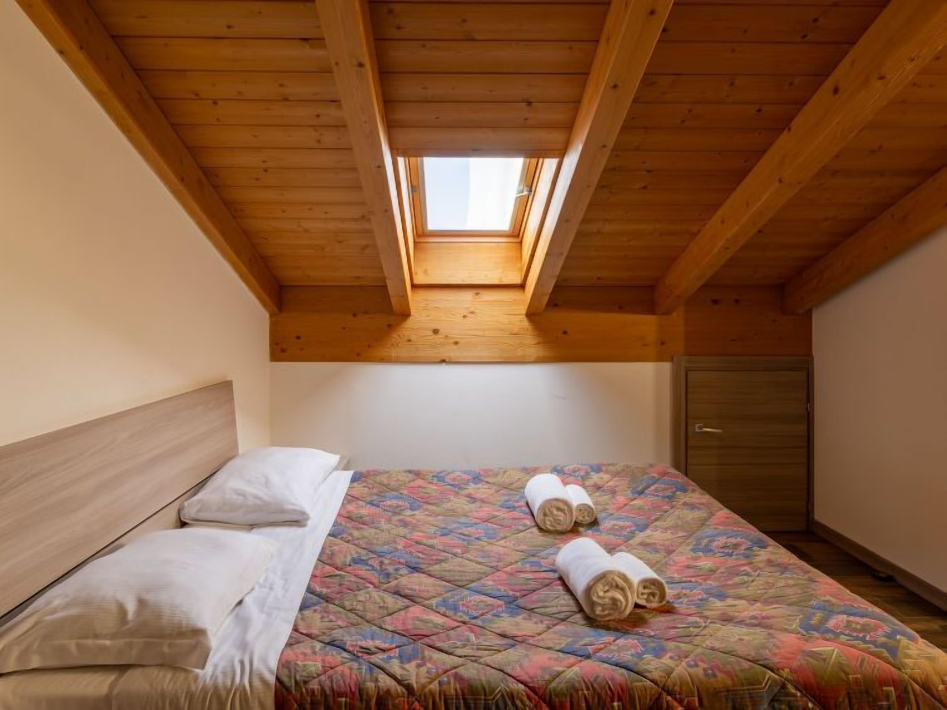 Un dormitorio con techo de madera y tragaluces, una cama con colcha de colores, almohadas y toallas enrolladas.