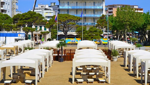 Un hôtel en bord de mer, avec de nombreuses tentes blanches et des chaises longues, un grand bâtiment doté de balcons, et un ciel d’un bleu limpide.