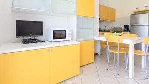 Une cuisine moderne avec des meubles de couleur jaune, un plan de travail blanc, un four à micro-ondes, un téléviseur à écran plat et un coin repas avec des chaises jaunes.