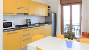 Une cuisine de taille réduite avec des meubles de couleur jaune, un plan de travail blanc, une cuisinière, un four à micro-ondes et un réfrigérateur. On y trouve une table à manger avec des chaises et une plante en pot.
