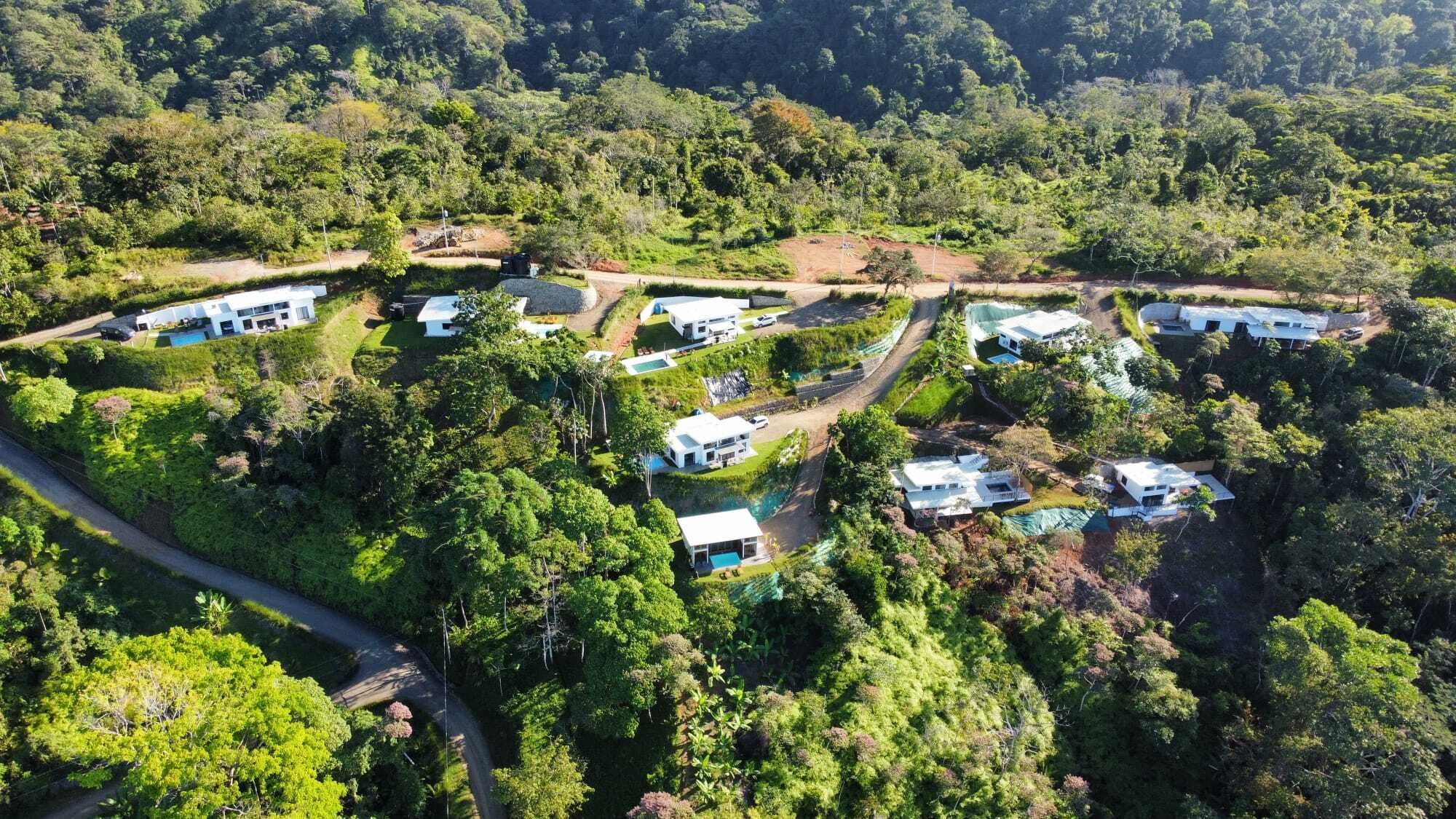 An aerial view of a residential area with multiple modern houses, surrounded by dense greenery and hills.