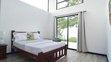 A bedroom with a wooden bed, a nightstand, a large window with curtains, and a view of greenery outside.