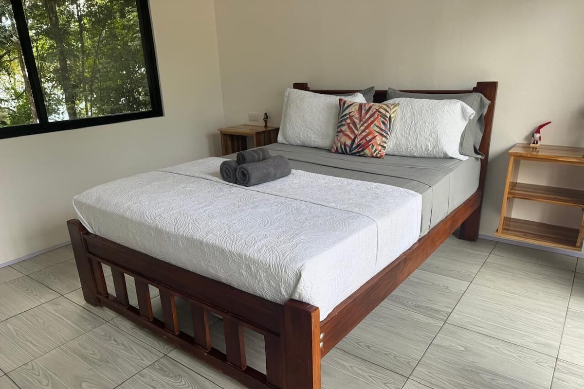 A double bed with a wooden frame, a grey mattress, and a white bedspread. There is a nightstand with a lamp and a small table beside the bed.