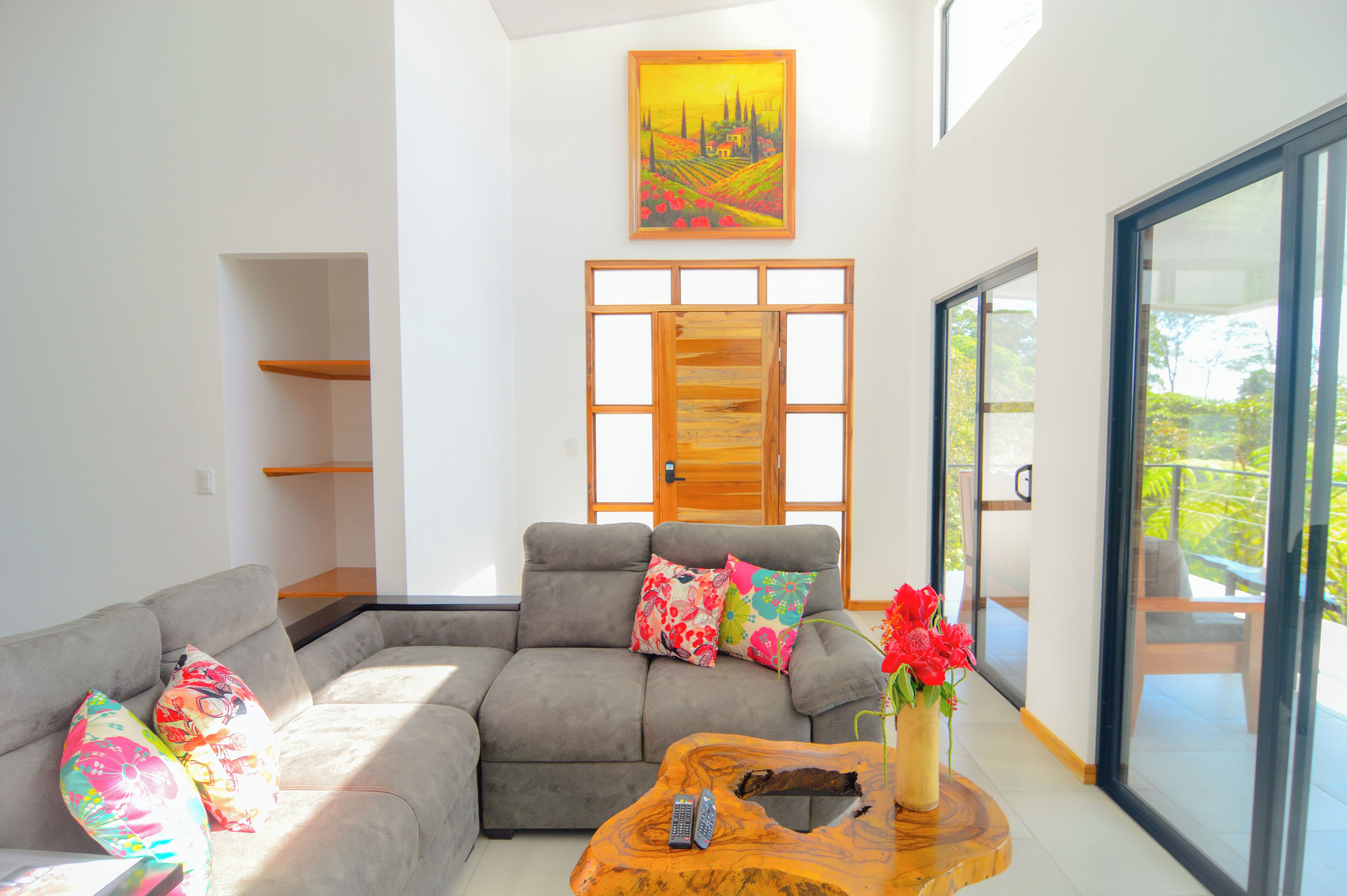 Una sala de estar moderna con un sofá seccional gris, almohadones coloridos, una mesa de centro de madera con un jarrón de flores rojas y un amplio ventanal con vistas a la vegetación.