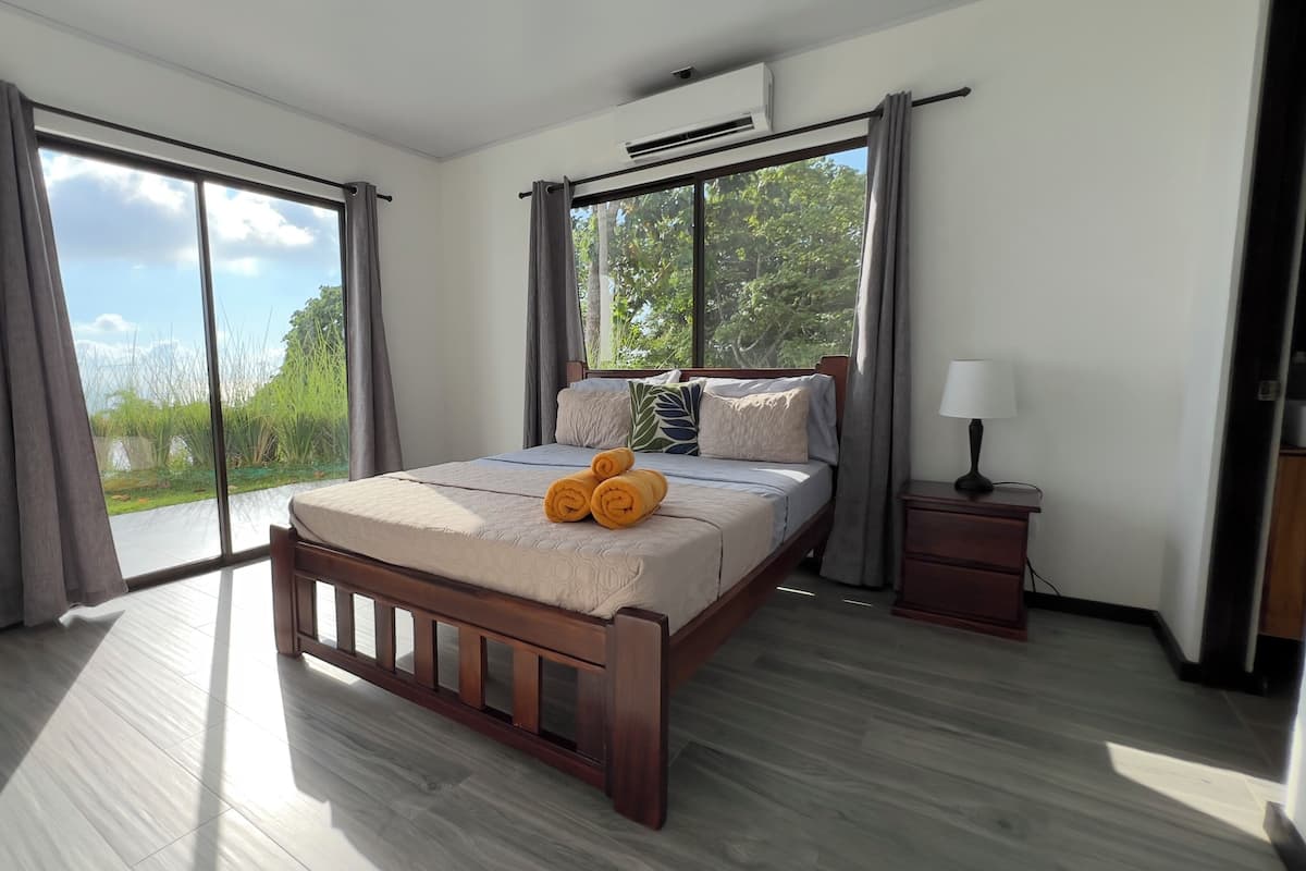 A bedroom with a large bed, a nightstand, an air conditioning unit, and a view of greenery through the window.