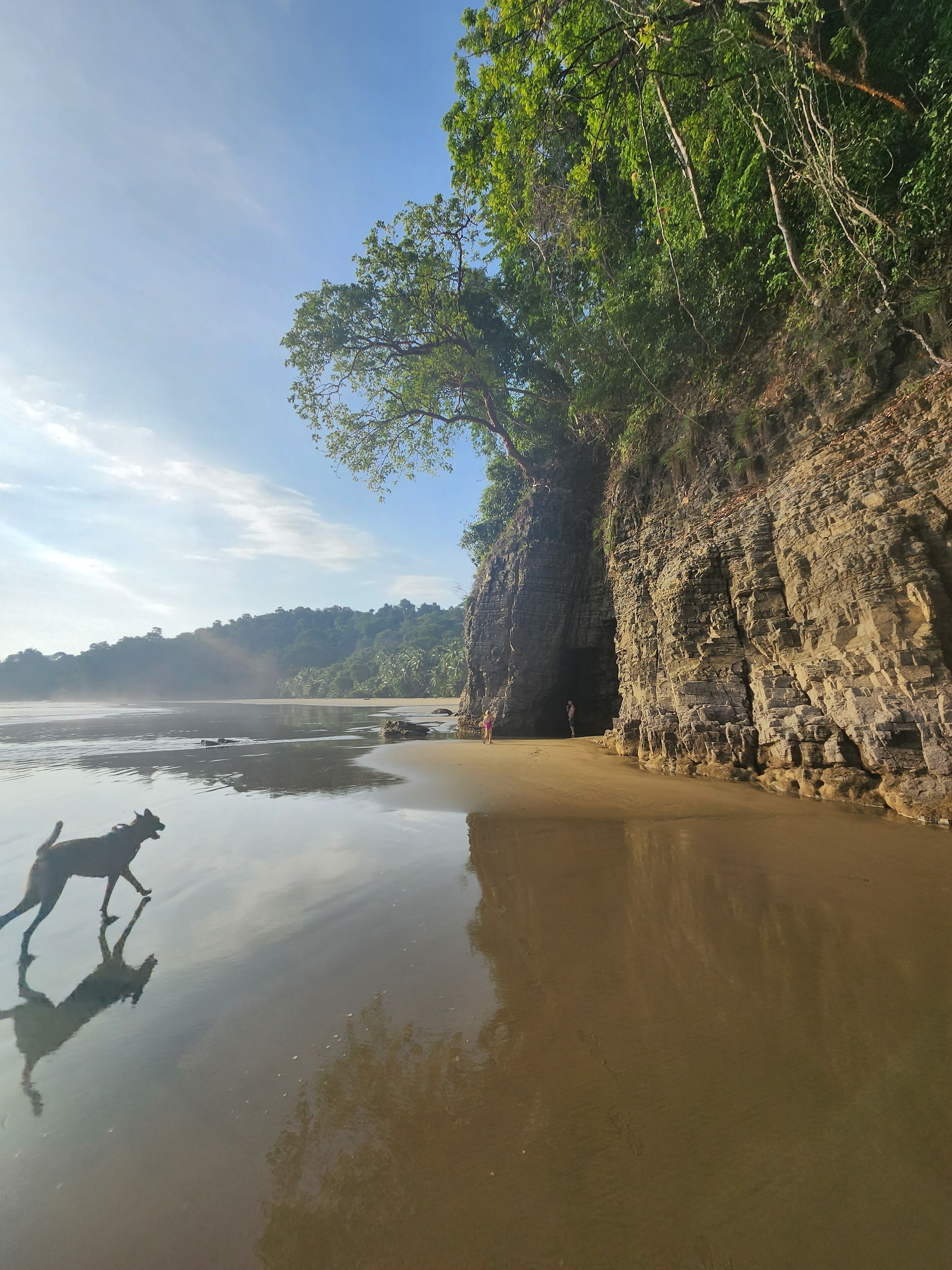 Una playa con un acantilado rocoso y una superficie de agua tranquila que refleja el cielo.