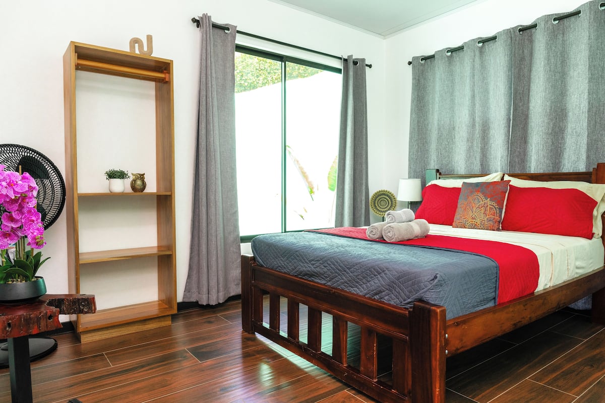 A bedroom with a wooden bed, a bedside table with a fan, a shelf with decorative items, and a window with curtains.