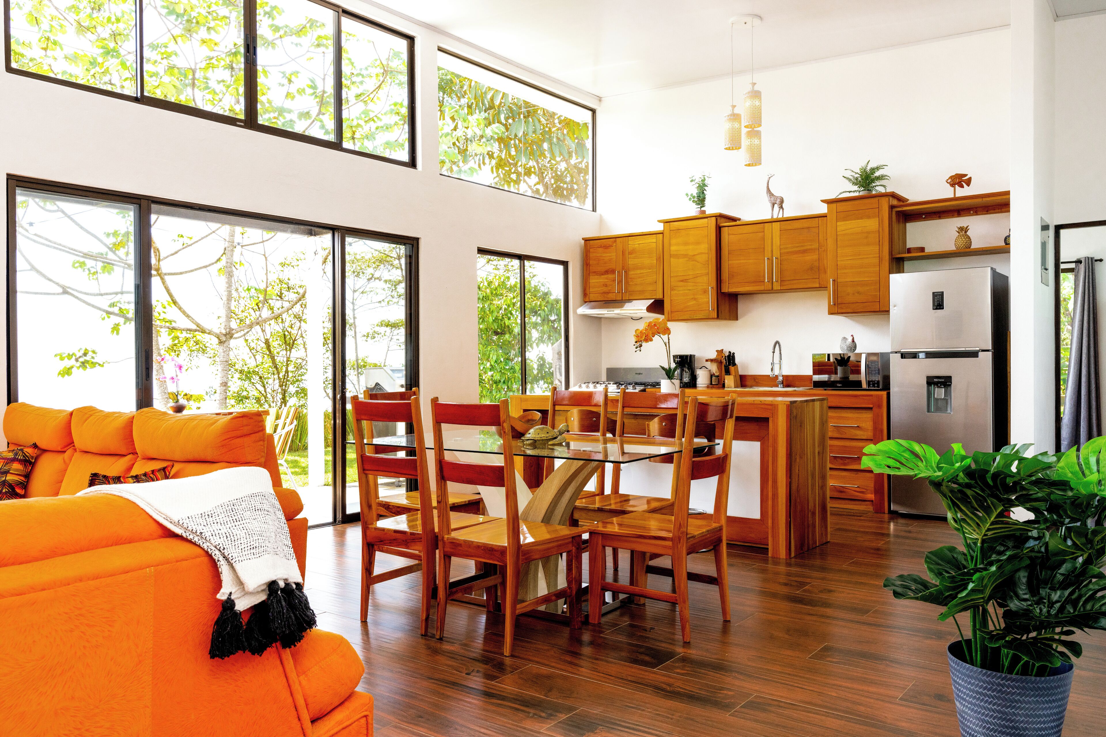 A modern kitchen with wooden cabinets, a dining area with orange furniture, and large windows offering views of greenery.