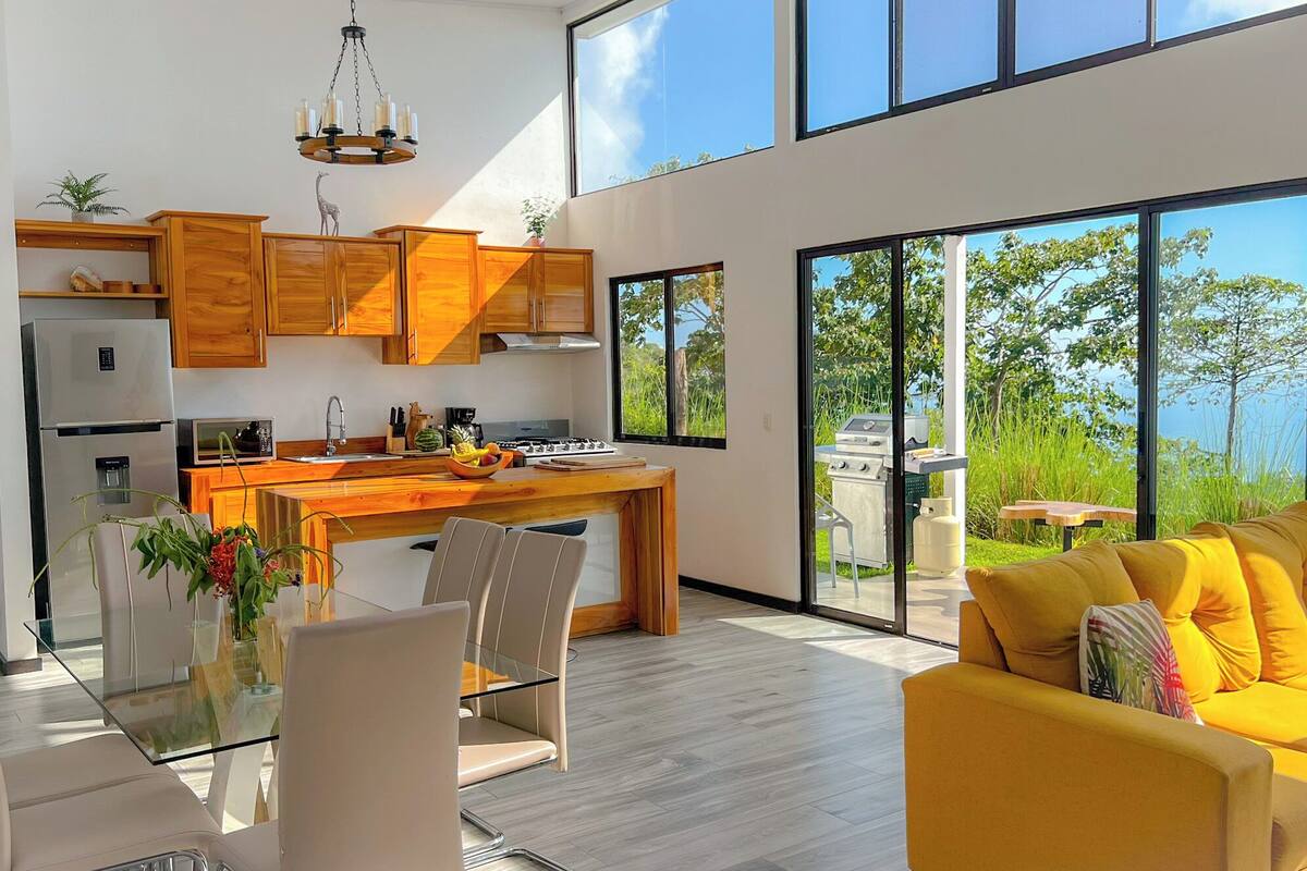 A modern kitchen with wooden cabinets, a dining area with a glass table, and a yellow sofa.