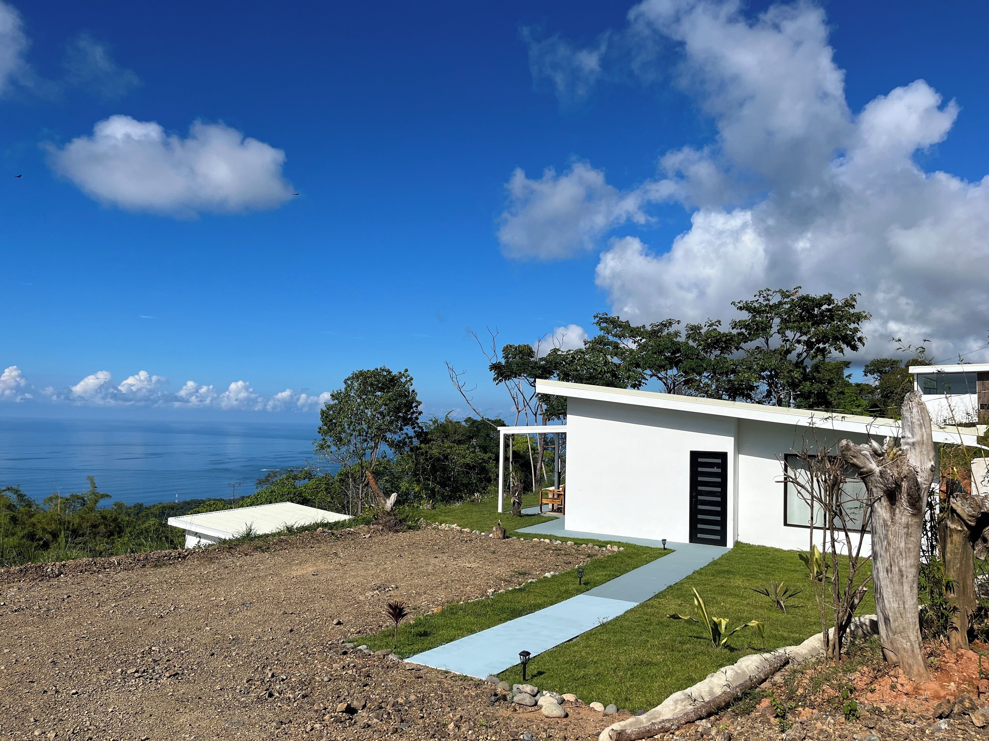 Une maison moderne aux murs blancs, avec une porte noire, un chemin pavé et une vue sur l’océan.