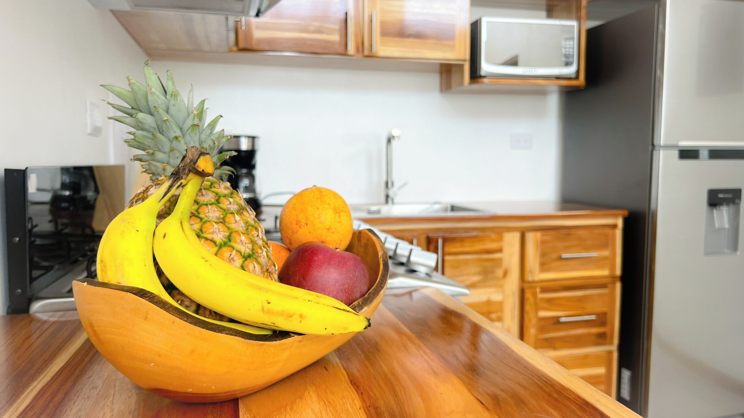 Une cuisine avec des meubles de rangement en bois, un four à micro-ondes et un panier de fruits contenant des bananes, un ananas et une pomme, le tout posé sur le comptoir.