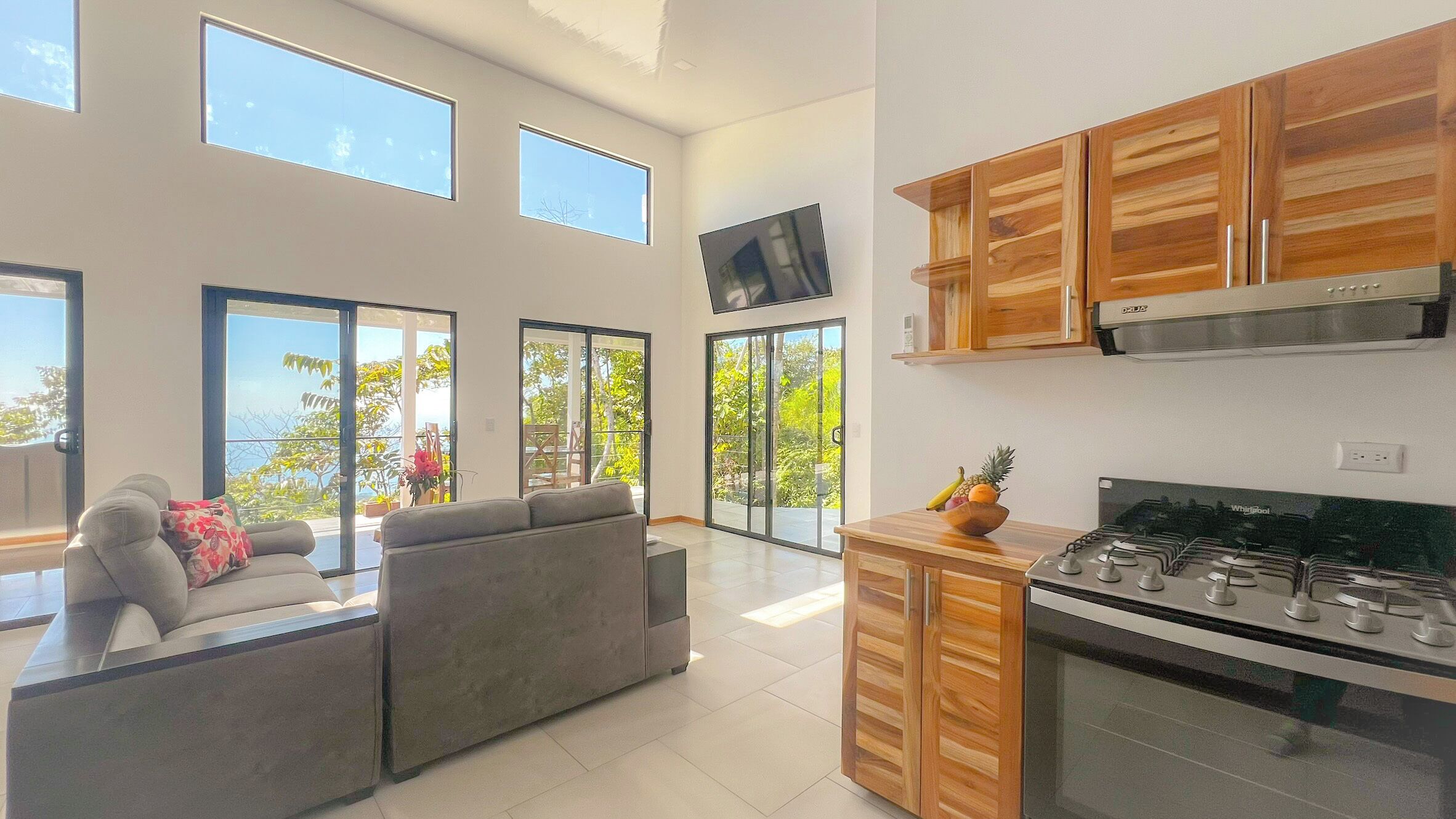 A modern kitchen with wooden cabinets, a stove, and a dining area with a sofa and a TV mounted on the wall.