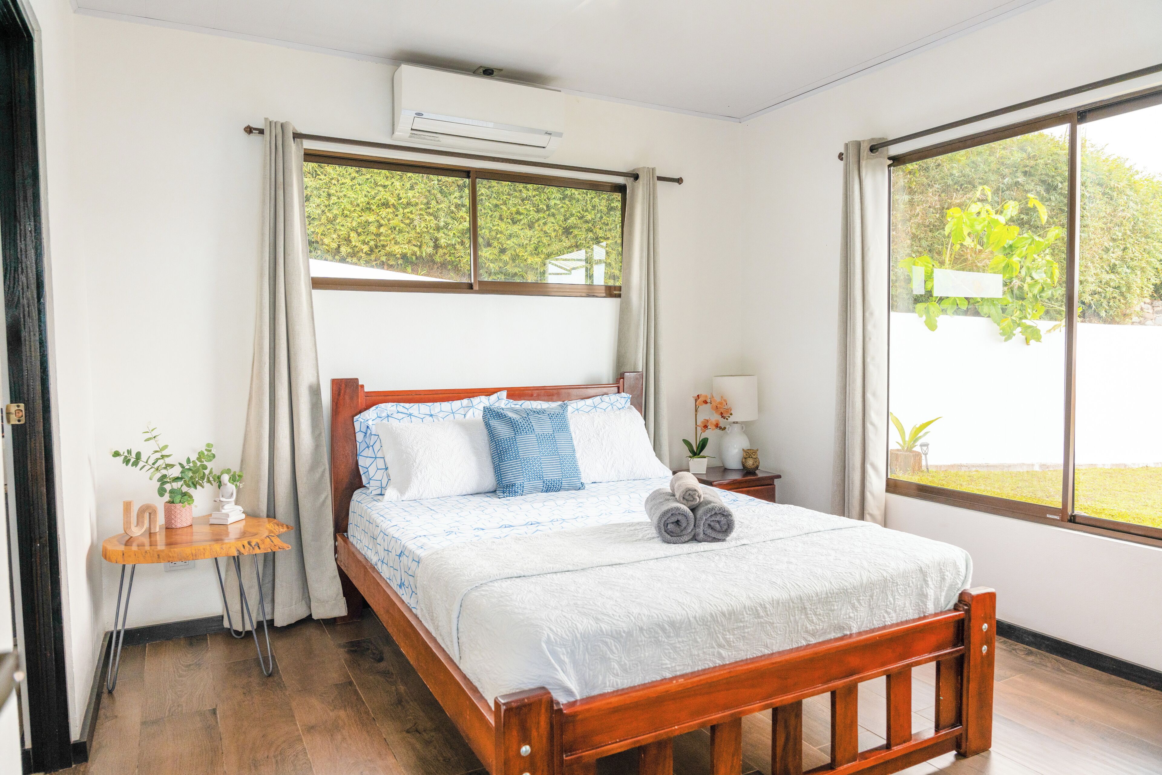 A neatly made bed with a wooden frame, a small bedside table with a plant, and a window with curtains.