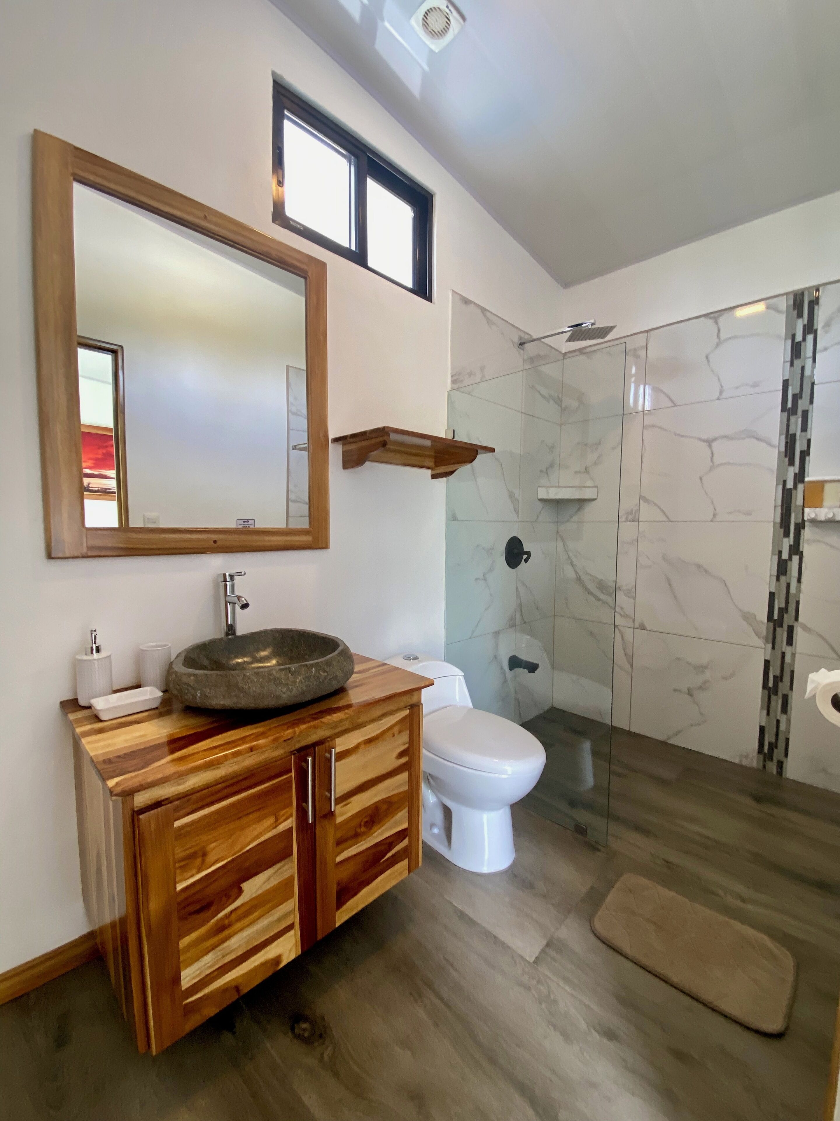A bathroom with a wooden vanity, a large mirror, a stone sink, a toilet, and a glass shower enclosure.