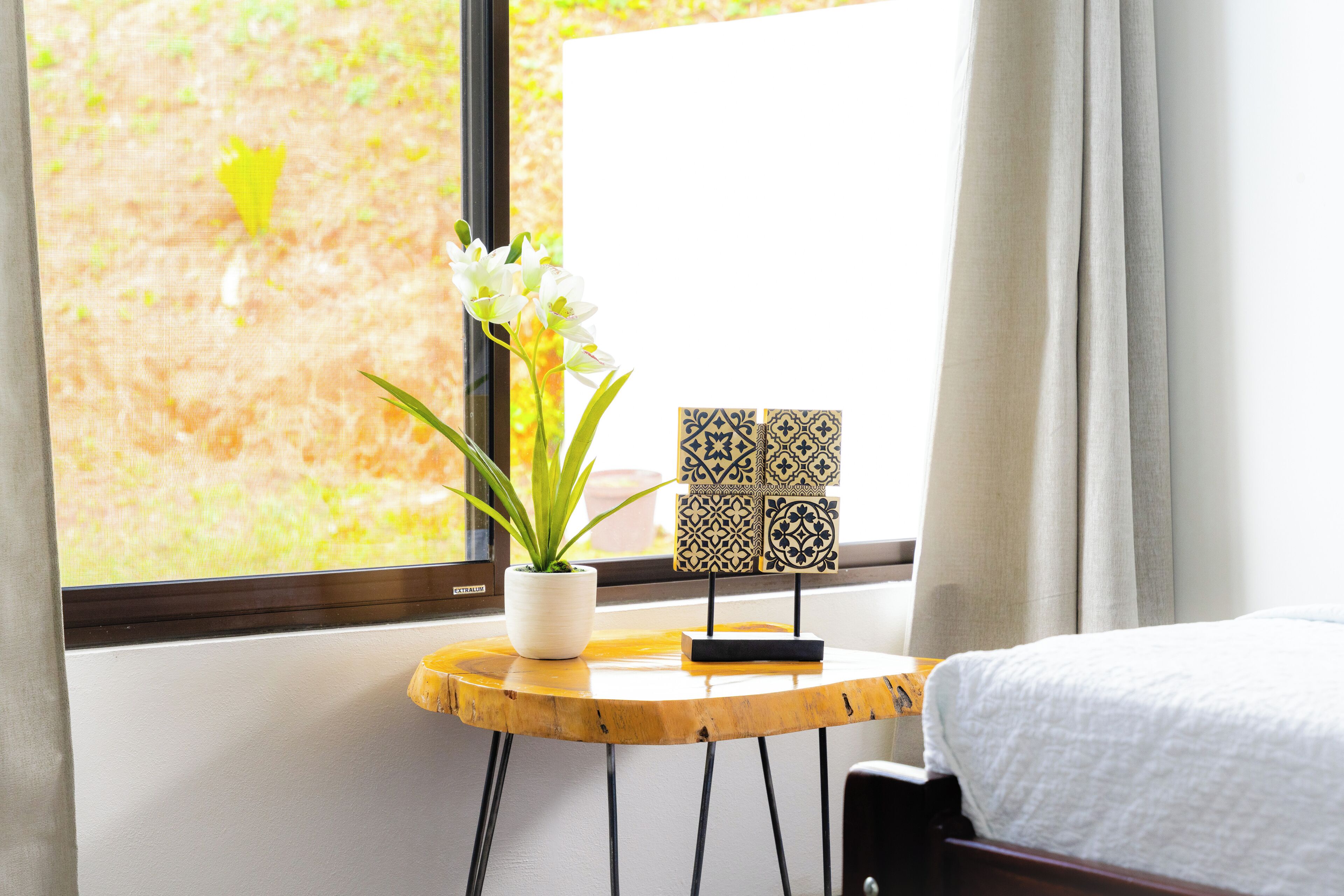 A hotel room with a bed, a small round wooden table with a potted plant and decorative tiles, and a window with a view of greenery.