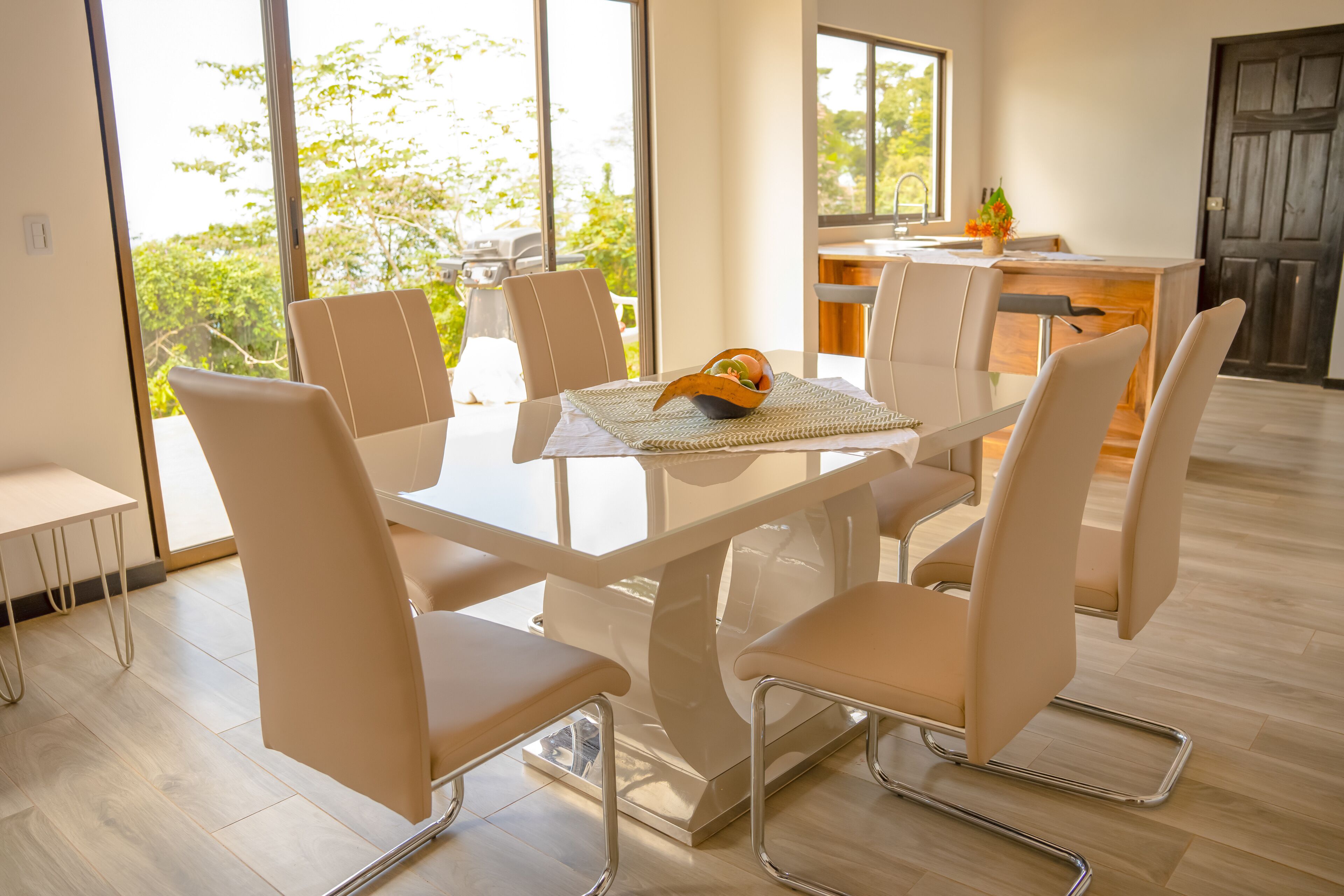 Uma área de refeições moderna com mesa de vidro e cadeiras brancas, cozinha ao fundo e grandes janelas com vista para o verde.