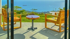Vista desde un balcón con dos sillas de madera y una mesita redonda, que da a un paisaje costero con vegetación y el mar.