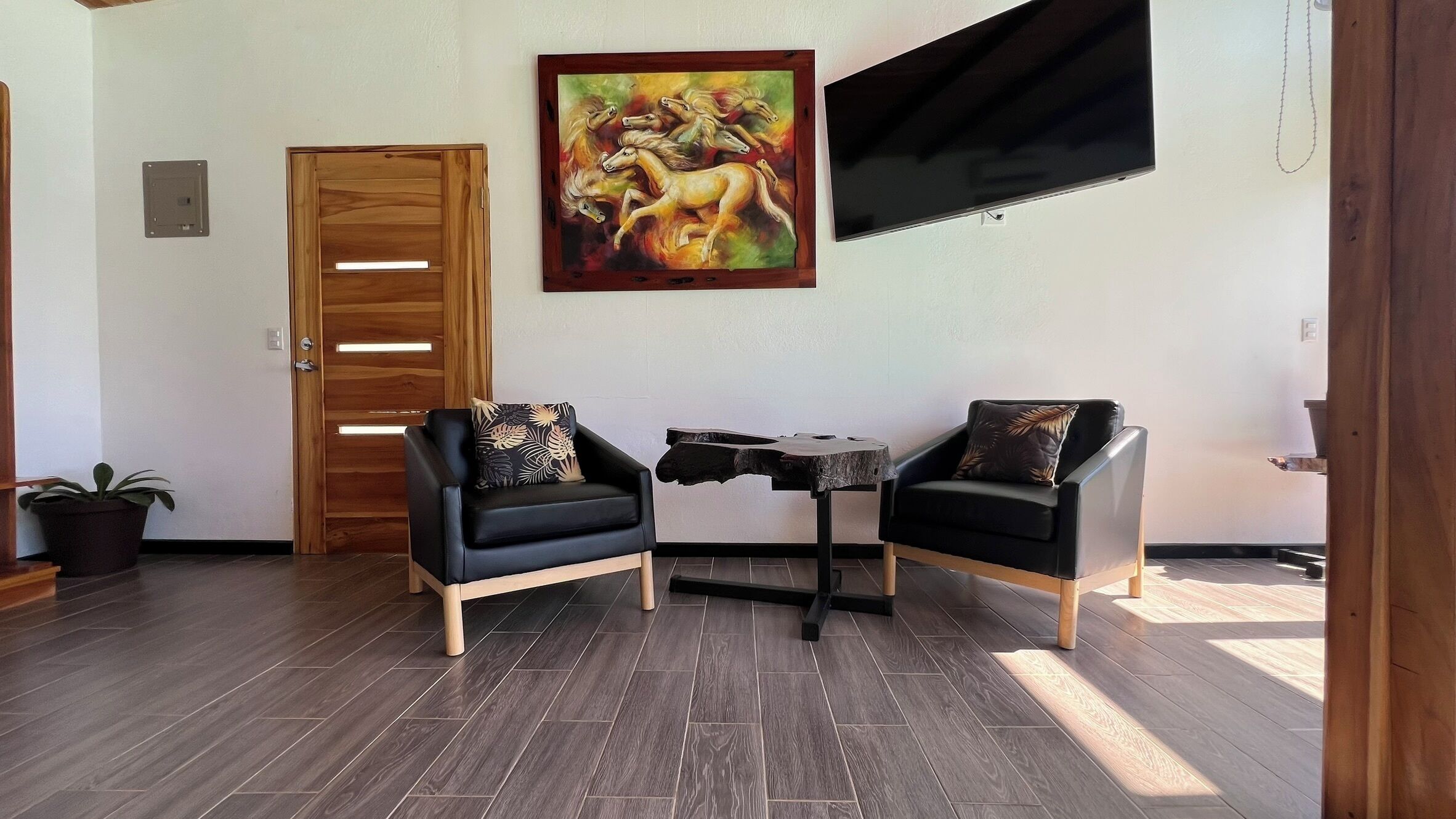 A room with two black armchairs, a small table, a painting of horses, a wooden door, and a TV mounted on the wall.