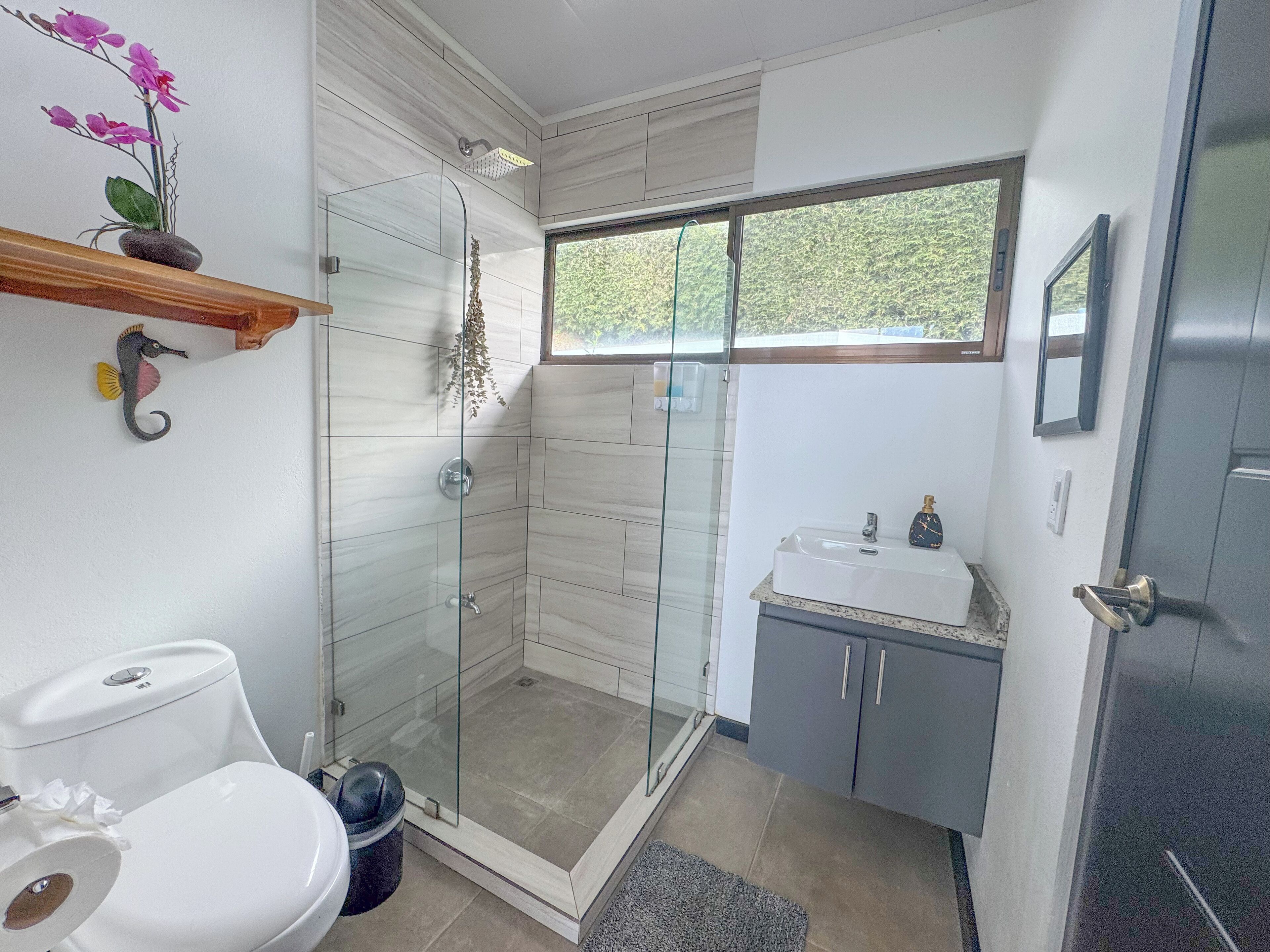 Une salle de bain moderne dotée d’une douche à parois de verre, de toilettes, d’un lavabo et d’une fenêtre laissant entrer la lumière naturelle.