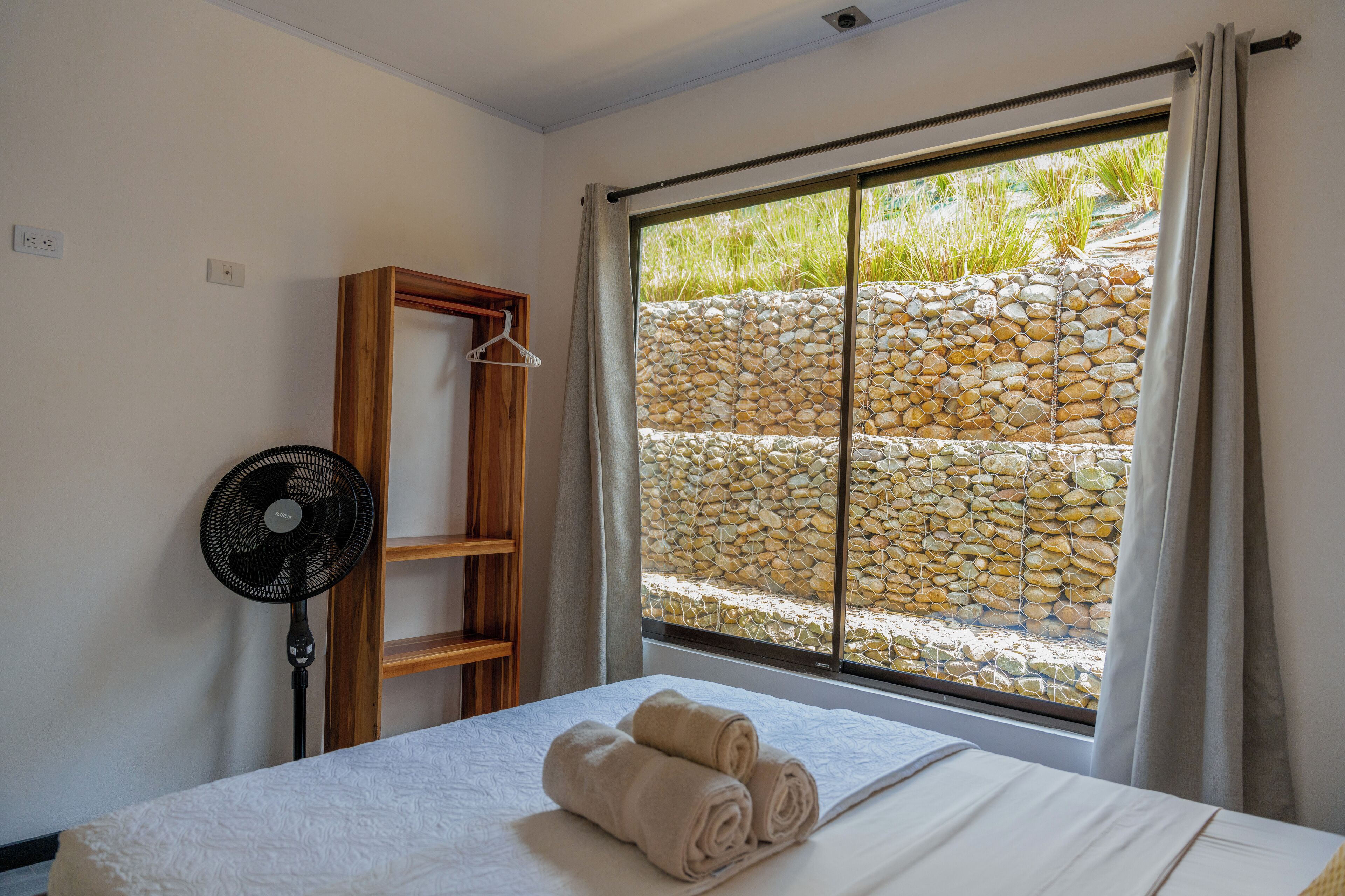 A hotel room with a bed, a fan, a wooden shelf, and a window with a stone wall view.