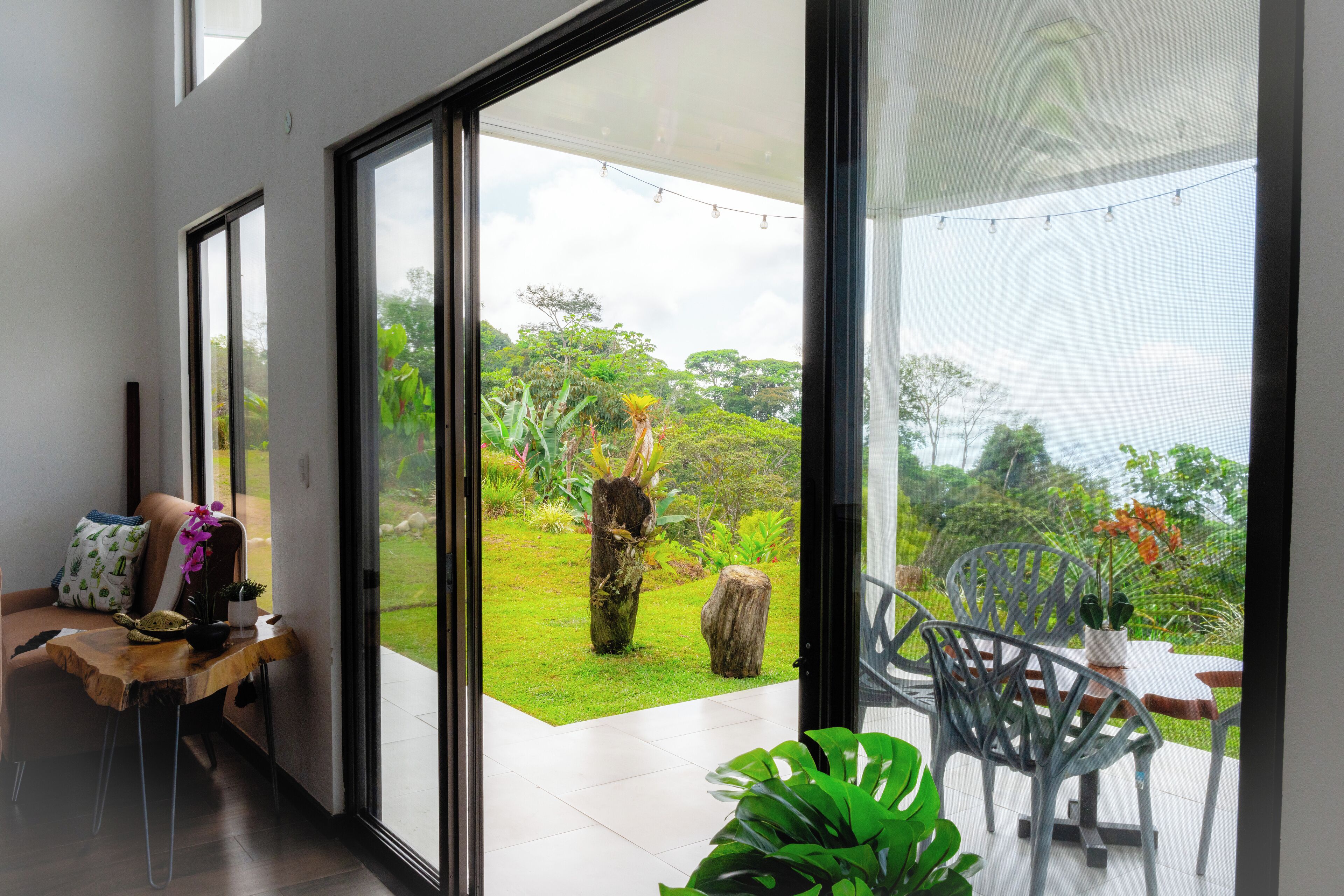 A modern room with a sliding glass door leading to a garden. Inside, there is a wooden bench with a potted plant and a small table. Outside, there is a dining area with chairs and a table, surrounded by greenery.