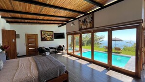 Un dormitorio con una cama grande, techo de madera, un cuadro, televisor y vistas a una piscina y jardín.
