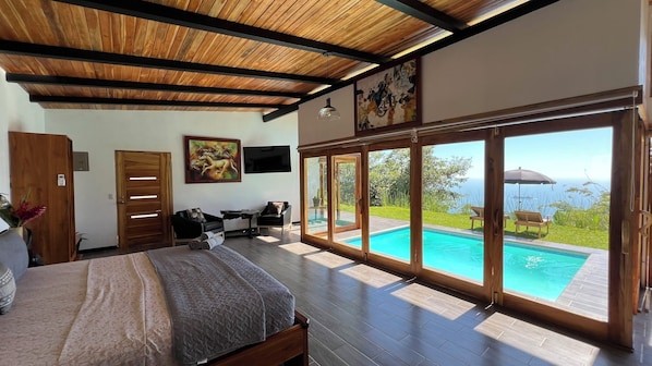 Un dormitorio con una cama grande, techo de madera, un cuadro, televisor y vistas a una piscina y jardín.